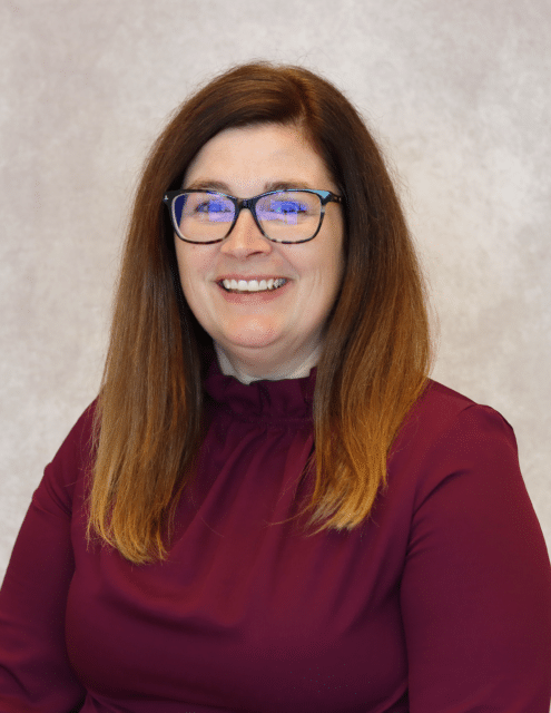 Sabrina Raja smiling, wearing glasses and a maroon blouse against a neutral background.